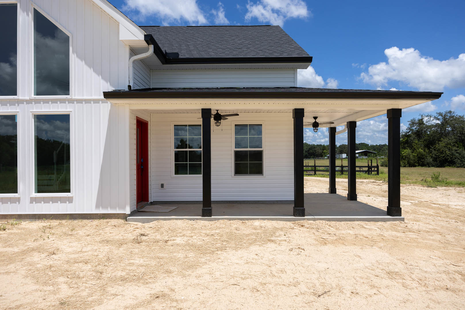 White siding exterior with red front door, large window reflecting nearby tree, dirt yard, porch roof detail, surrounding trees under cloudy sky