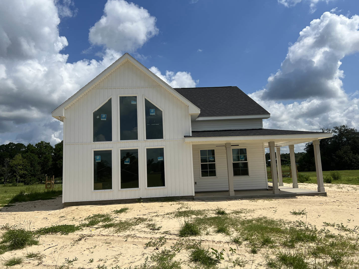 White two-story house with black roof, multiple windows, and front porch, set against blue sky with scattered clouds and surrounded by grass and trees.