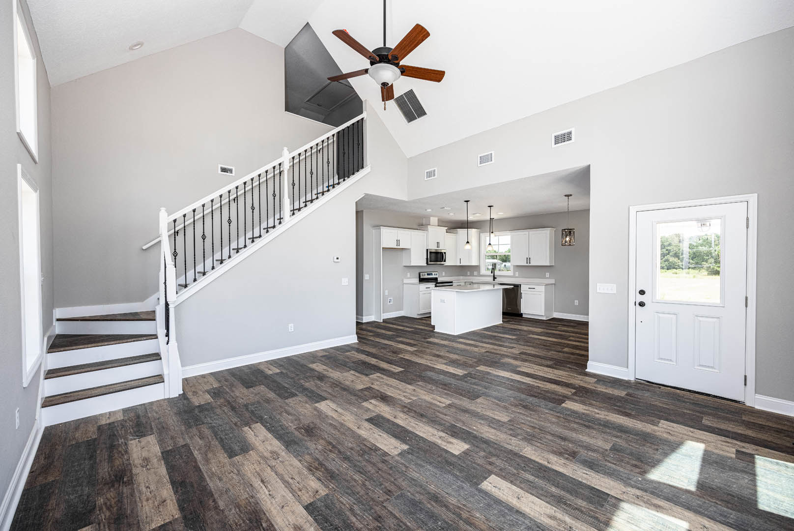 Open-concept kitchen with white cabinetry, black and white countertops, wood flooring, adjacent staircase featuring white railing, ceiling fan with light fixture, white door with