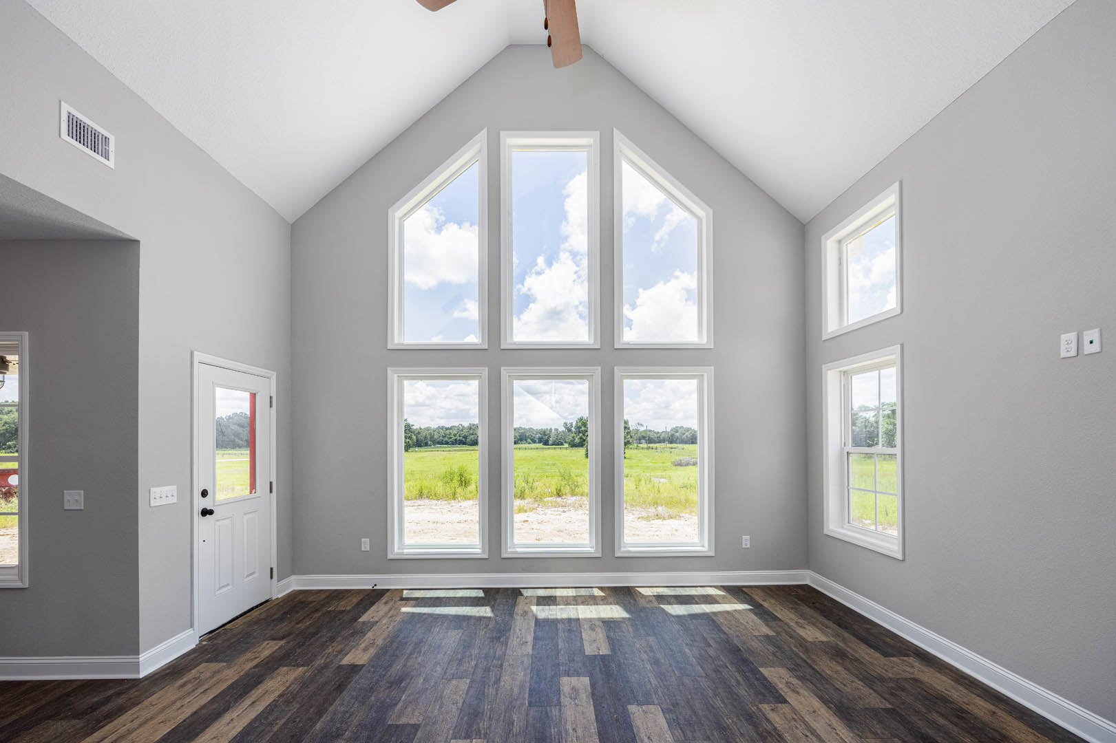 Ceiling fan mounted above wooden laminate flooring, multiple windows with views of grassy field, trees, and blue sky, white door with glass panel, natural light streaming into
