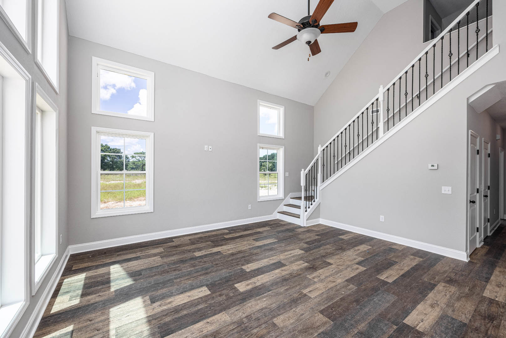 Open living area with wood flooring, white walls, ceiling fan with light fixture, staircase with wooden steps and railing, large window overlooking grassy field and blue sky