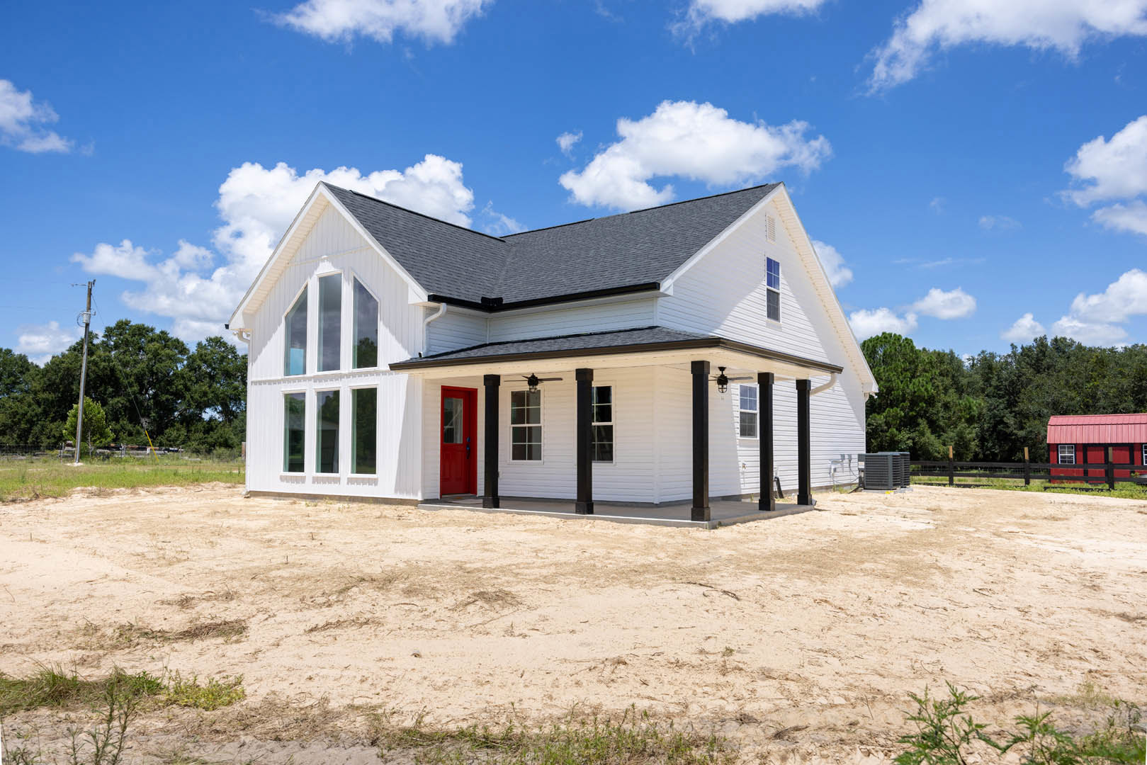 White siding house with a red front door, small porch, patchy grass and dirt yard, blue sky with clouds, nearby building and black fence, tree and street sign visible