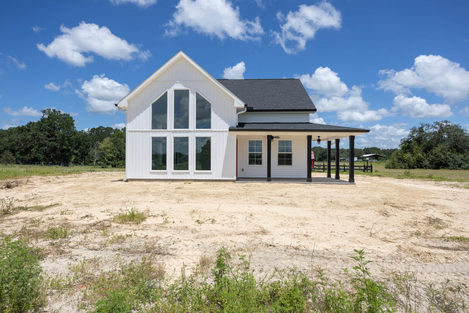 White siding house with black shingle roof, multiple white-framed windows, covered front porch, dirt yard with scattered plants, cloudy sky overhead