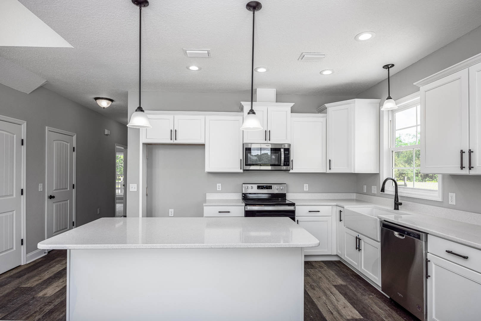 White kitchen with shaker cabinets, central white island, quartz countertops, stainless oven with open door, black hardware, ceiling light fixture, window showing blurred trees.