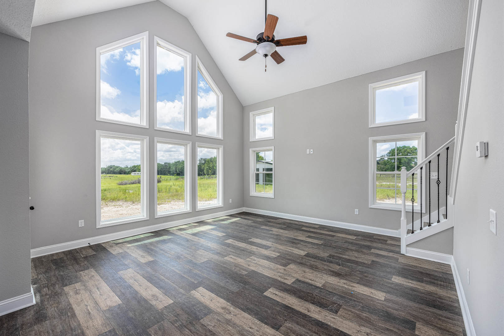 Ceiling fan with light fixture above wood flooring, white-framed windows showing blue sky, staircase with wooden steps, neutral walls