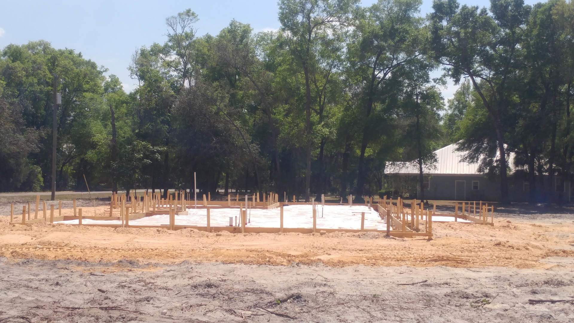 Wood-framed home under construction with exposed foundation, sandy ground, wooden deck, and fence, surrounded by tall trees in the background