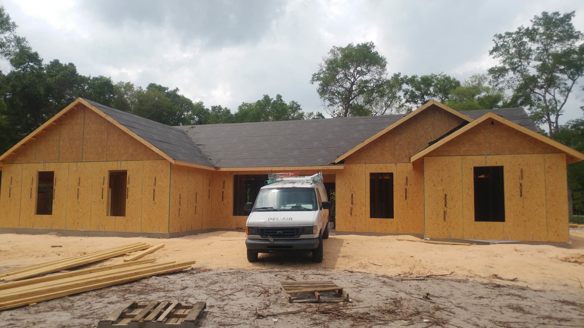 White van with roof light parked on sandy driveway in front of modern house with large windows, light-colored exterior, and surrounding trees.
