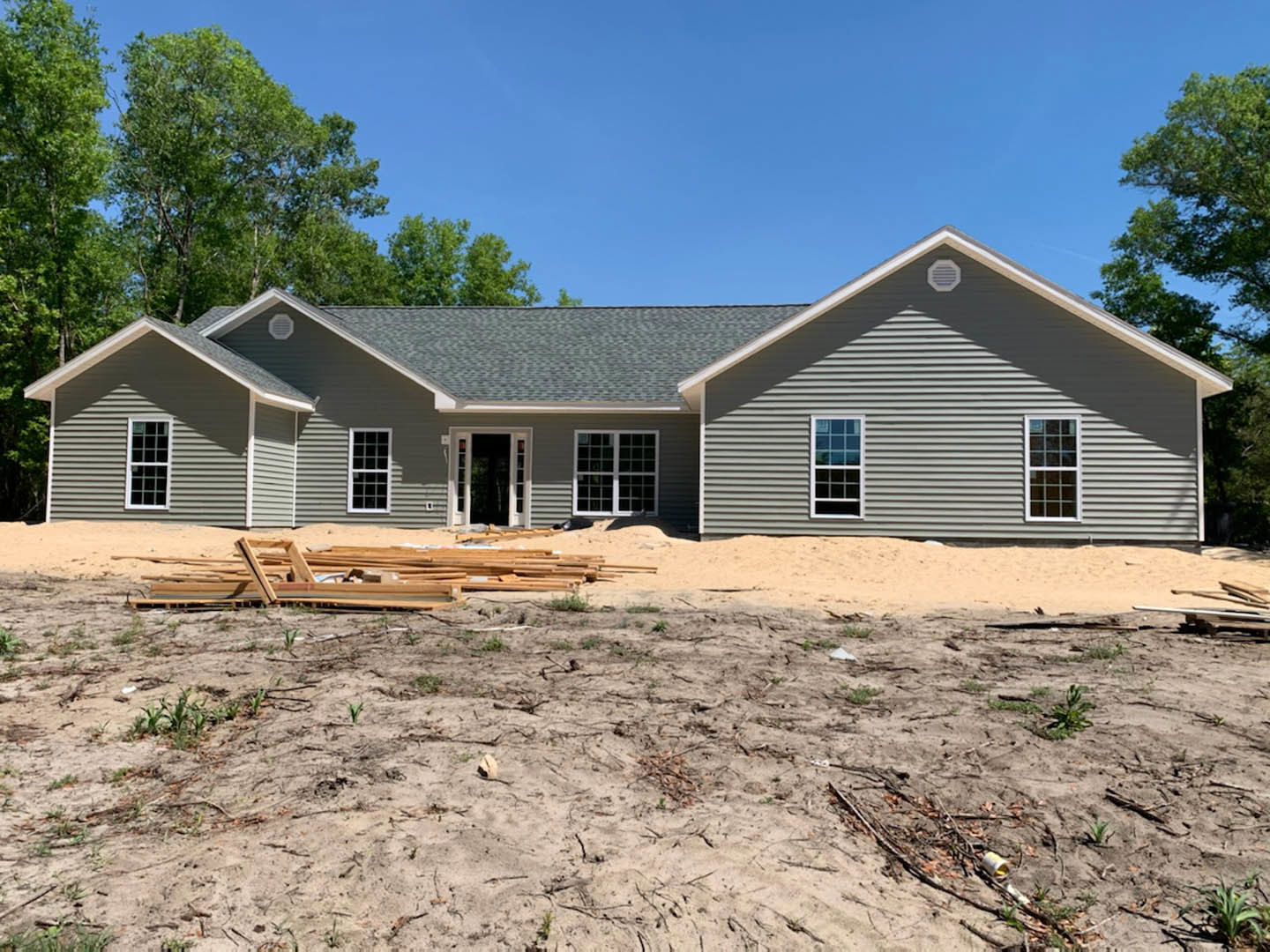 Partially built house with exposed framing, sandy ground, surrounding trees, multiple pane windows, and a white door with glass panels under a clear blue sky
