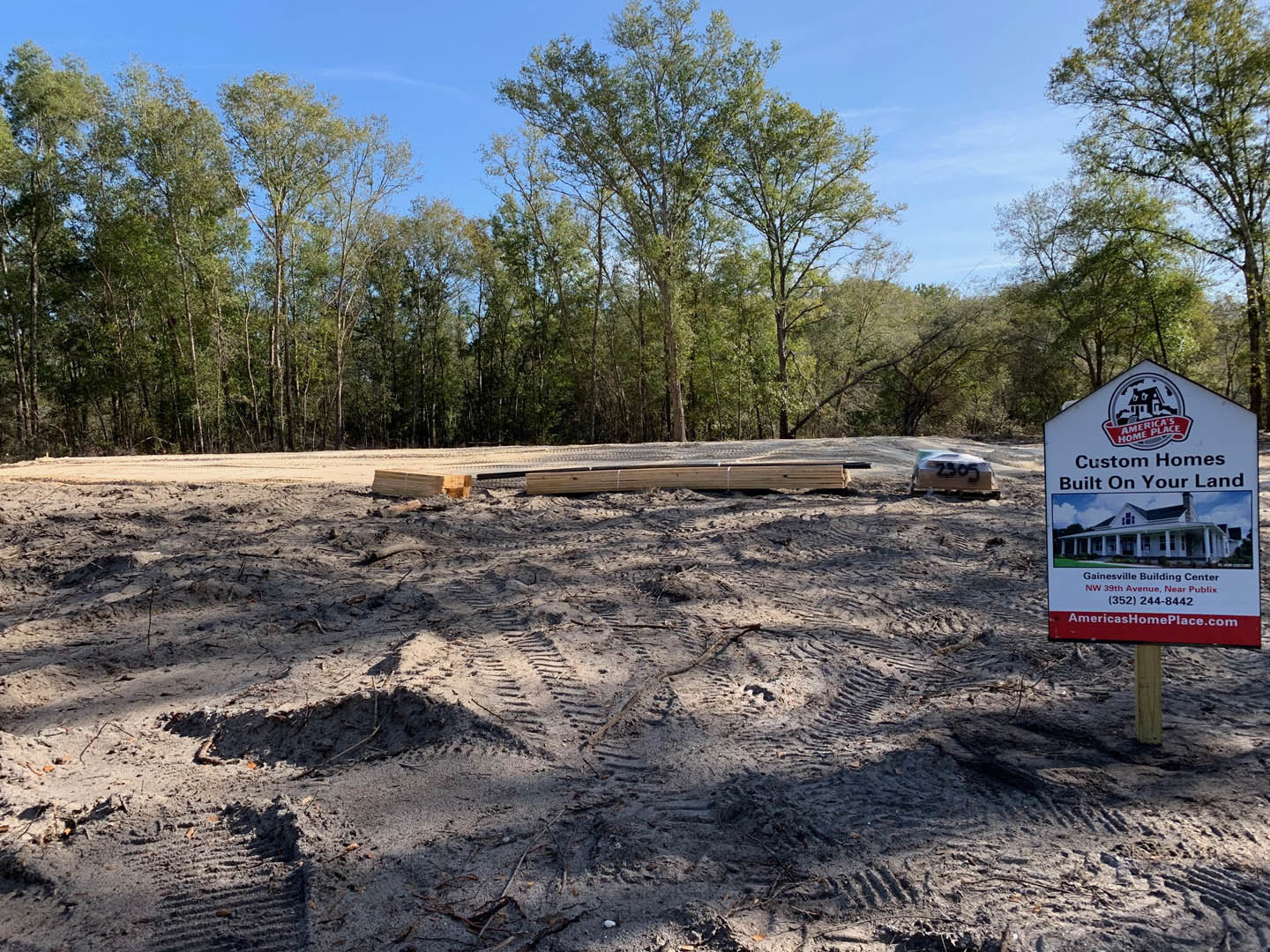 White sign with a house illustration and red ribbon stands in sandy soil with tire tracks, surrounded by trees and forested background; blurry car visible in distance.