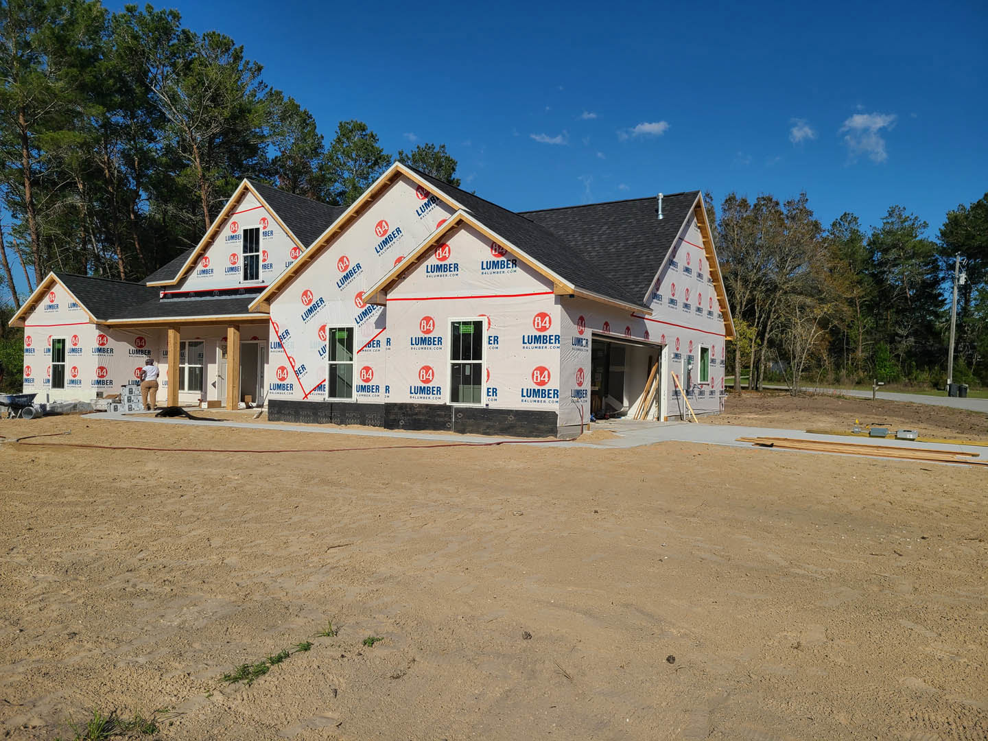 Partially built house with white roof covering, surrounded by dirt field, white-framed window visible, construction sign on roof, trees and cloudy sky in background