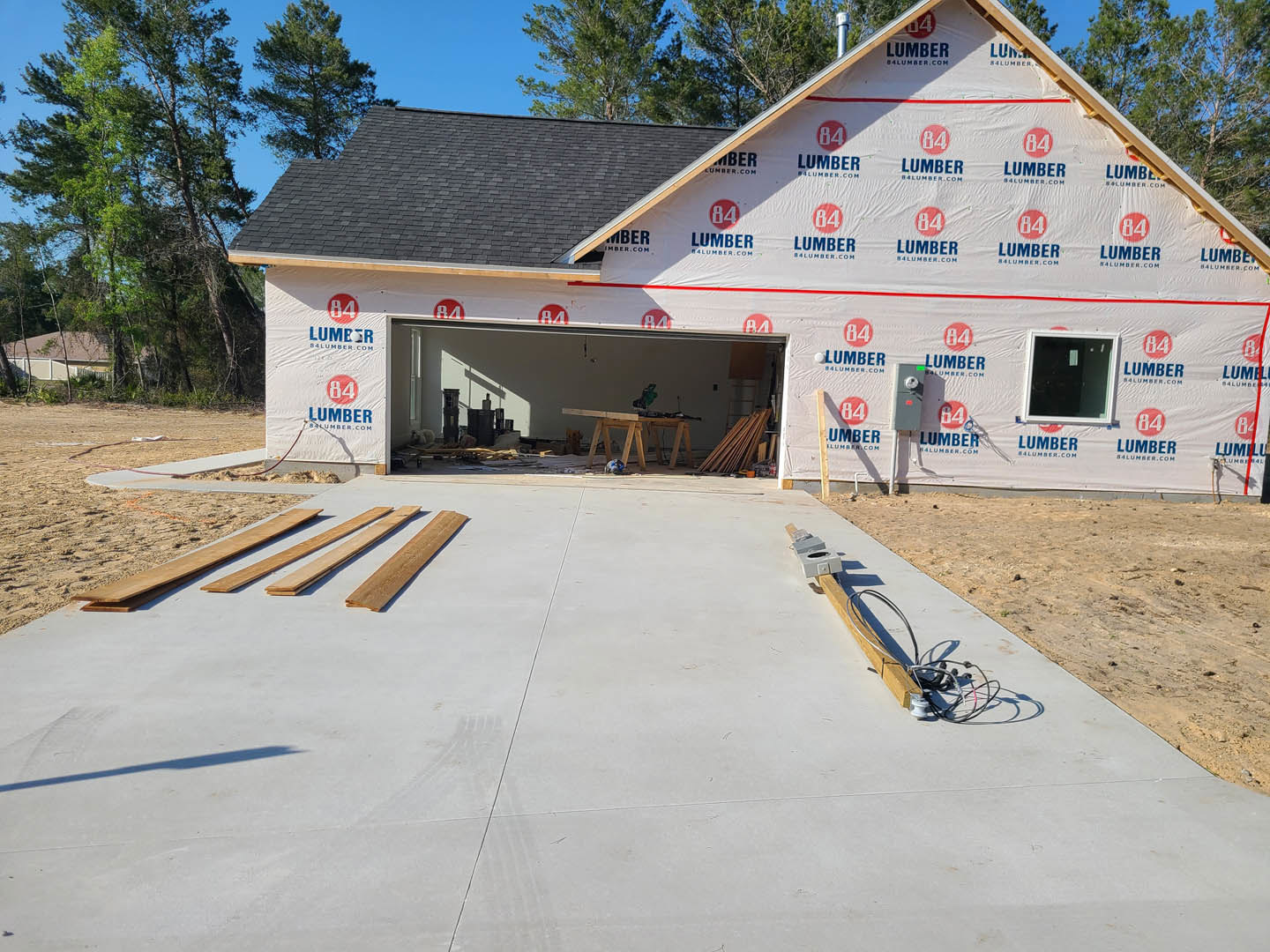 Partially built house with exposed wood framing, white-framed window, unfinished garage door opening, concrete floor with scattered wires and lumber, wood planks stacked on
