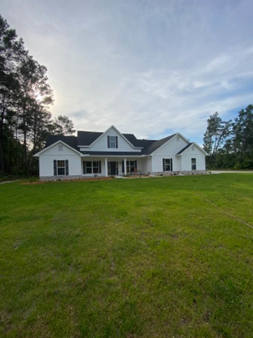 White two-story house with wide front porch, large green lawn, mature tree with sunlight filtering through branches, blue sky with scattered clouds