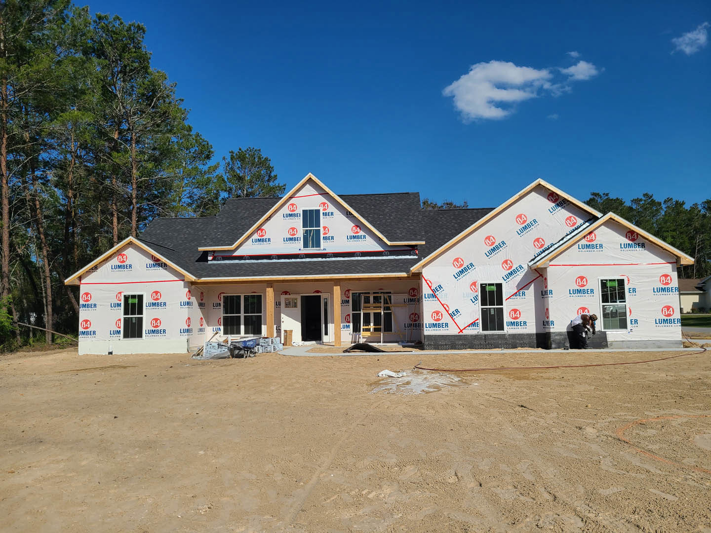Modern custom home under construction with exposed dirt lot, light-colored siding, large windows, and blue sky overhead; trees visible in background.