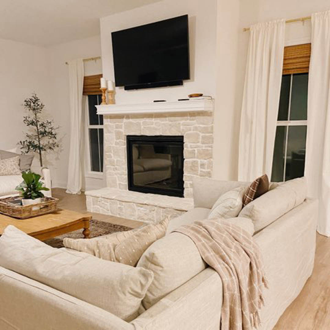 Living room with stone fireplace, gray upholstered couch draped with a blanket, wall-mounted black TV, potted green plant beside sofa, neutral walls, hardwood flooring