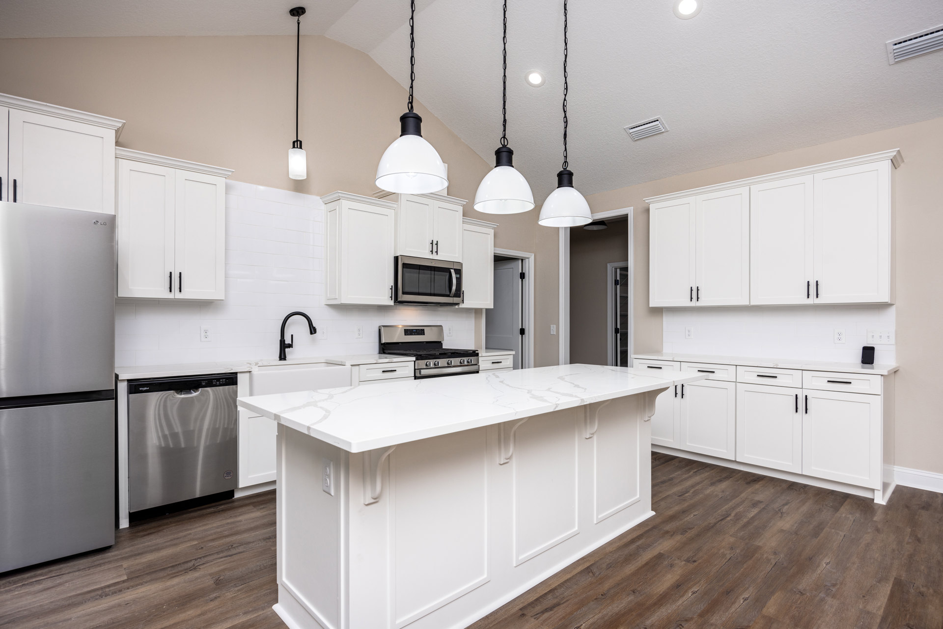 White cabinetry kitchen featuring a spacious island with built-in stove and oven, stainless steel refrigerator, open microwave, dishwasher with black handle, ceiling vent, and