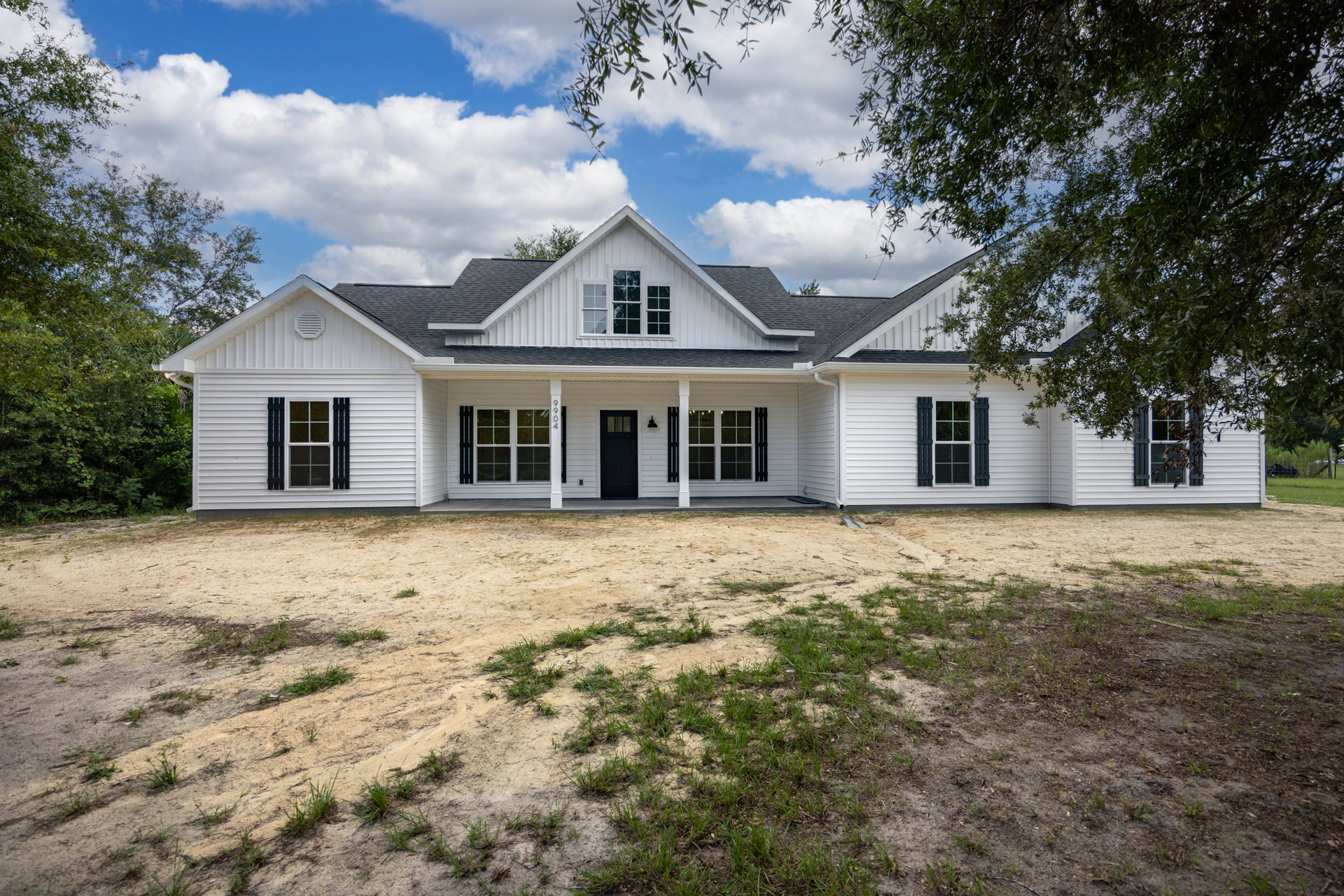 White farmhouse with black door and window shutters, dirt yard, and cloudy sky in background