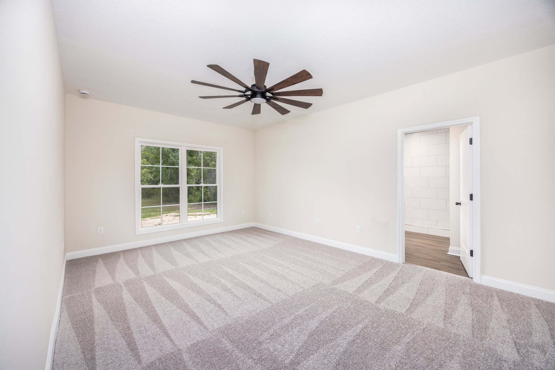 Carpeted bedroom with white walls, ceiling fan with light, large window showing trees, open white door leading to bathroom