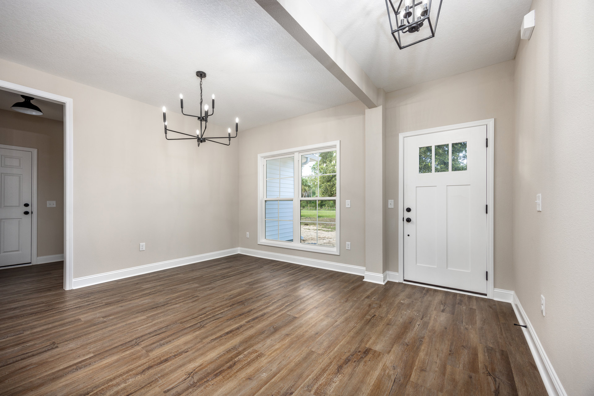 White paneled door with black knobs beside a large window, wood laminate flooring, white plaster walls, black metal chandelier with exposed bulbs hanging from ceiling