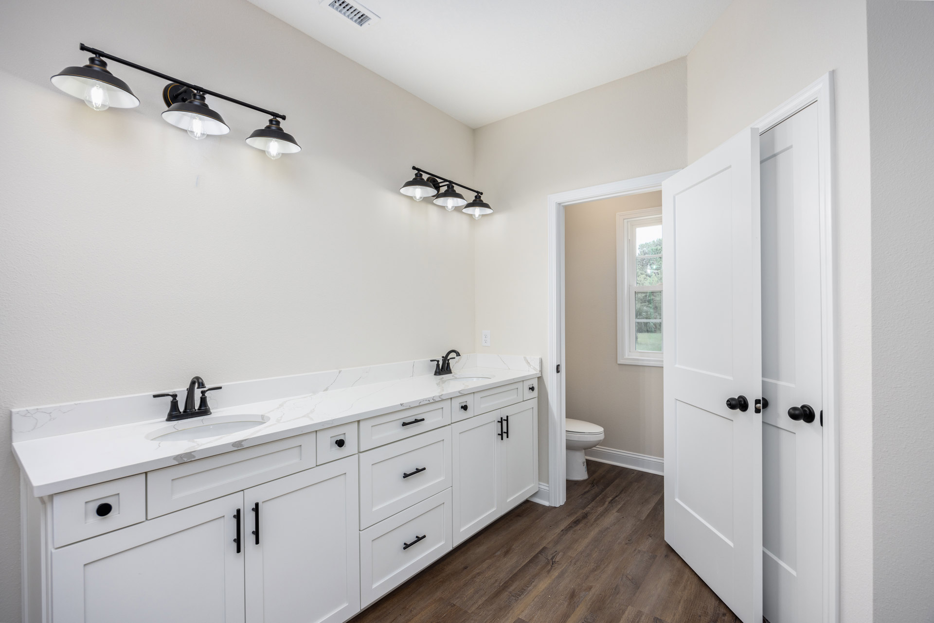 Bathroom with white shaker cabinets, black light fixtures, marble countertop, black faucet, and window above toilet.