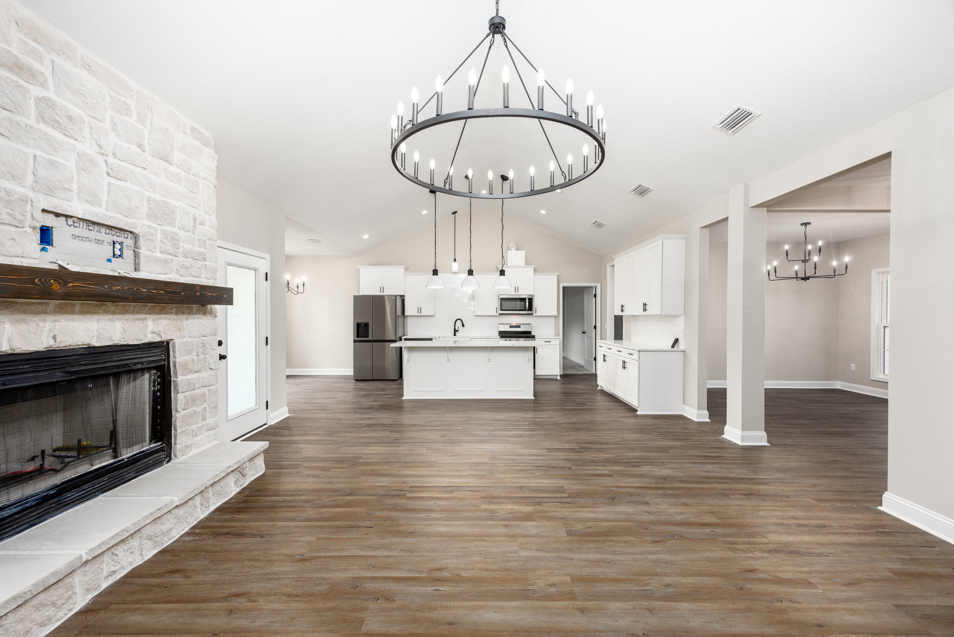 Open plan living room featuring wood flooring, central fireplace with metal screen, ceiling chandelier, white plaster walls with vent, glass panel door, and stainless steel