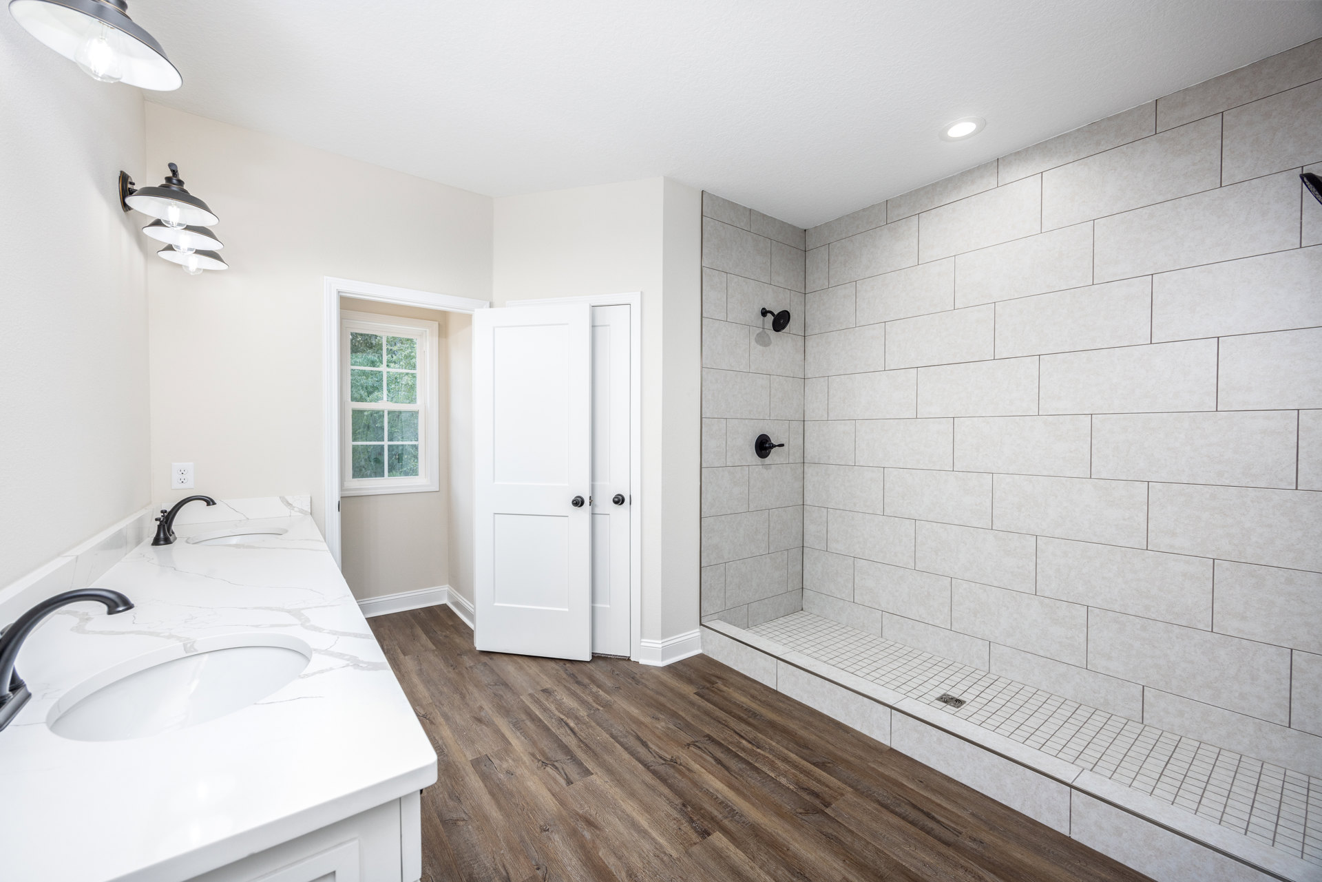 Bathroom with white countertop and black faucets, wall-mounted light fixture, window with white frame, and shower featuring tile floor.