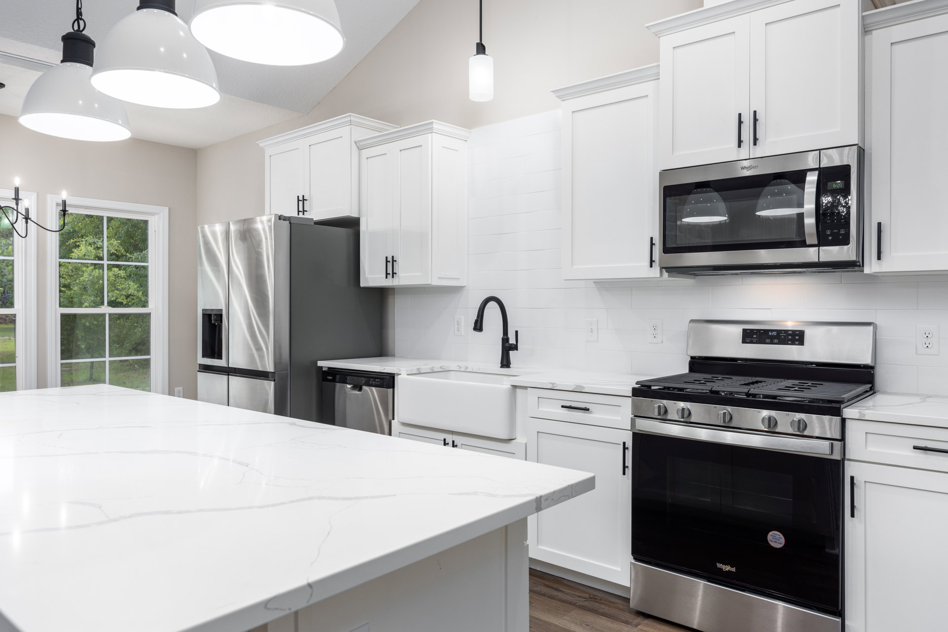 White kitchen with shaker cabinets, marble countertop, stainless steel appliances including stove, oven, and open refrigerator, built-in microwave, large window overlooking trees.