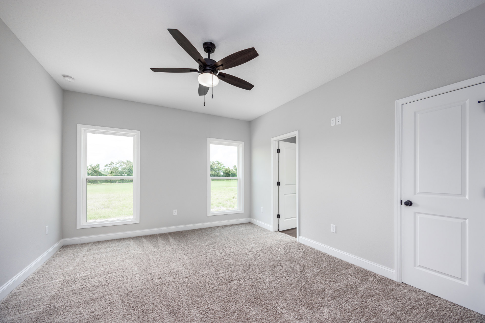 Neutral-toned room with wood flooring, white walls, ceiling fan with light, two windows showing grass and trees outside, white door with black hardware