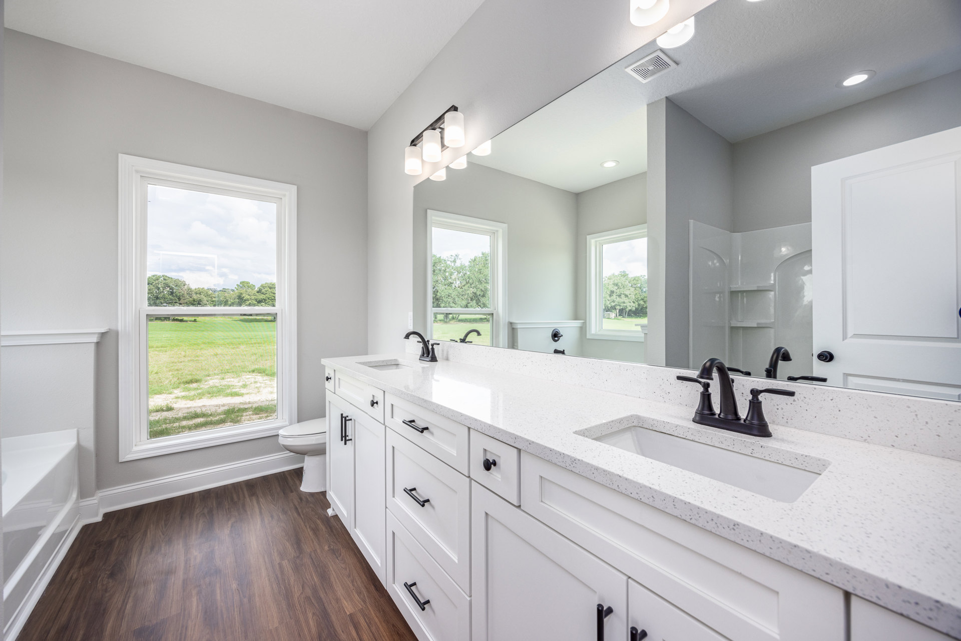 Bathroom with wood flooring, white trim, expansive mirror above a large sink, ceiling vent, and a wide window overlooking a grassy field.