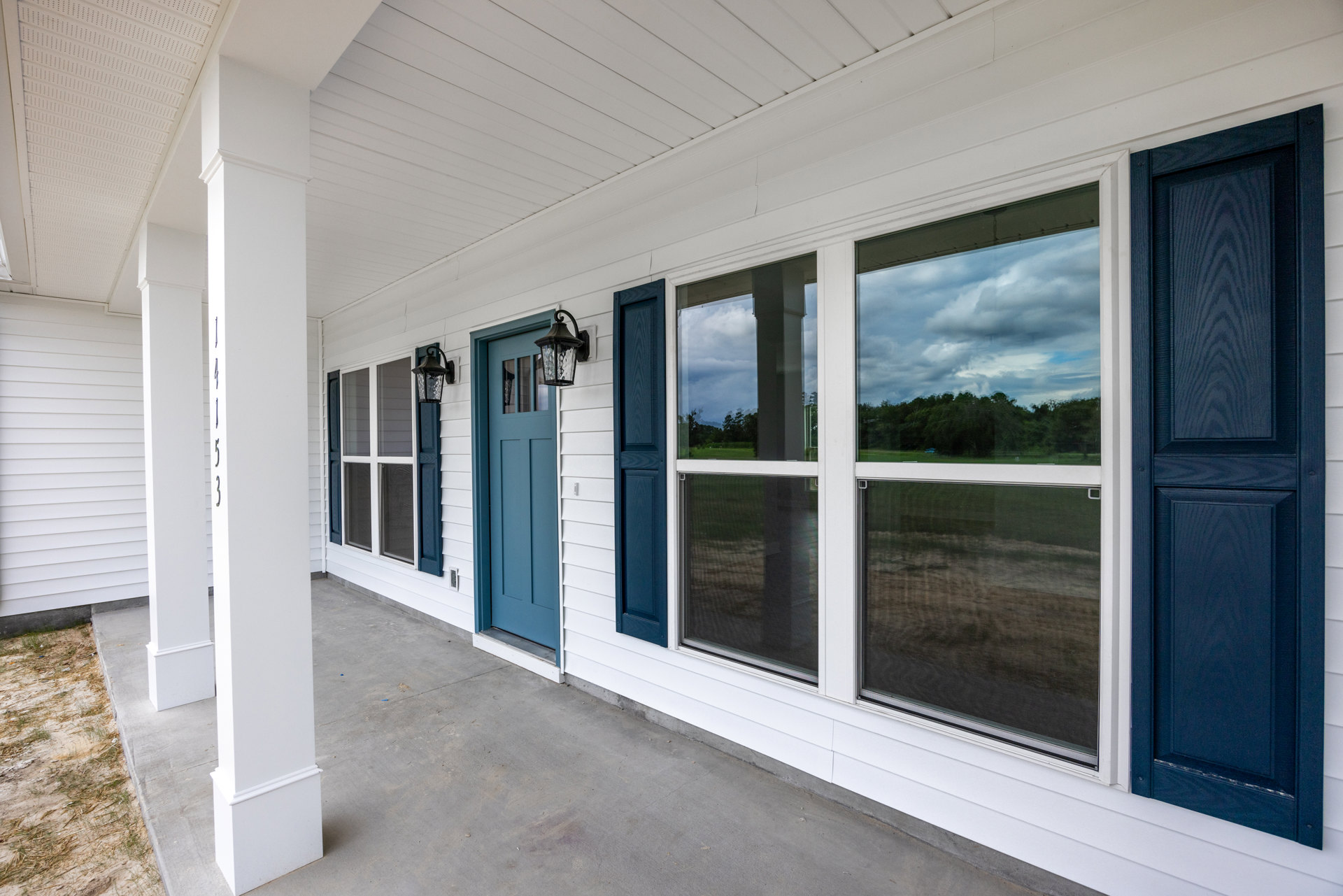 Front porch with white siding, blue door, blue-trimmed windows, outdoor lantern fixture, and view of trees reflected in window glass