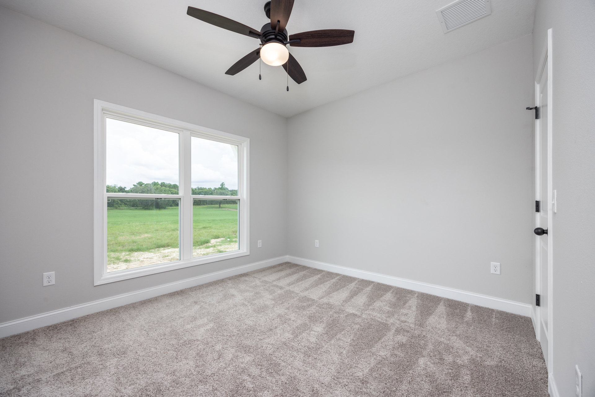 Ceiling fan with integrated light fixture mounted on white ceiling, large window overlooking grassy yard, beige carpet flooring, smooth plaster walls