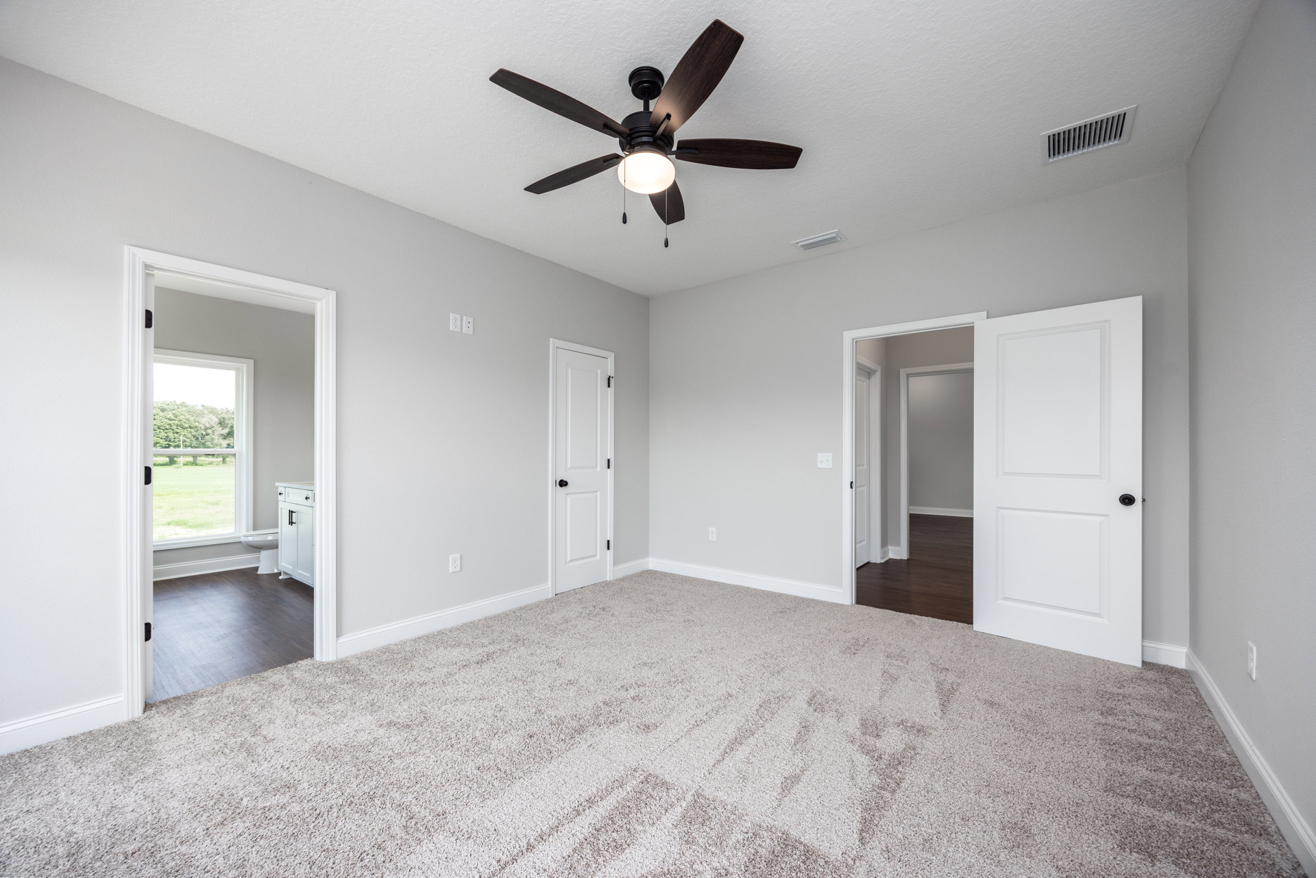 Carpeted room with white walls, ceiling fan with light fixture, white door with black knob, and wall vent