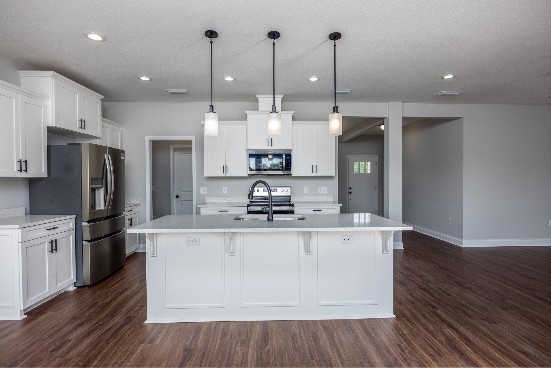 Spacious kitchen featuring a large central island with white cabinetry and black handles, stainless steel refrigerator, built-in sink and stove, wide window reflecting trees, light