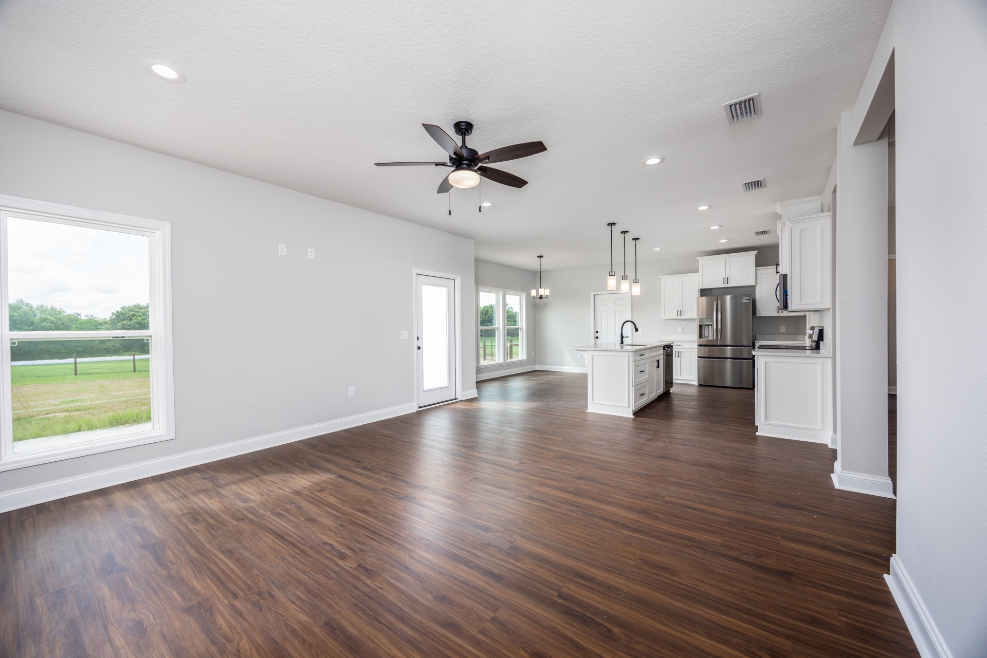 Spacious white room featuring wood flooring, ceiling fan with light, white kitchen island with black handles, refrigerator, and window overlooking fenced grassy yard