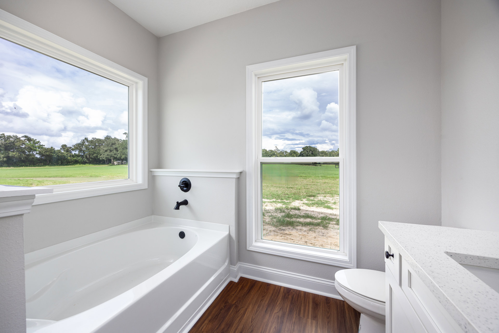White bathtub with black fixtures beside a window showing grass and trees, wood flooring with white trim, white countertop with black handle, neutral walls.