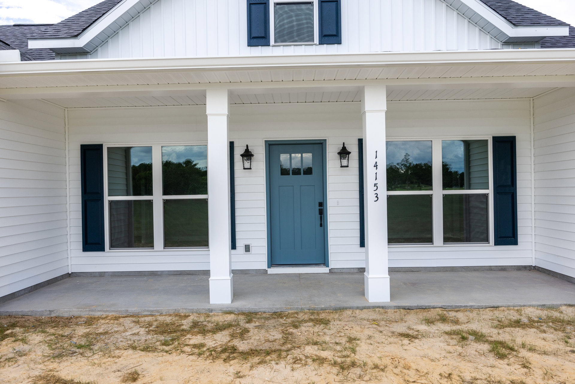 White siding house with blue front door and black handle, flanked by columns, screened window, patchy grass and dirt yard, white post displaying house numbers