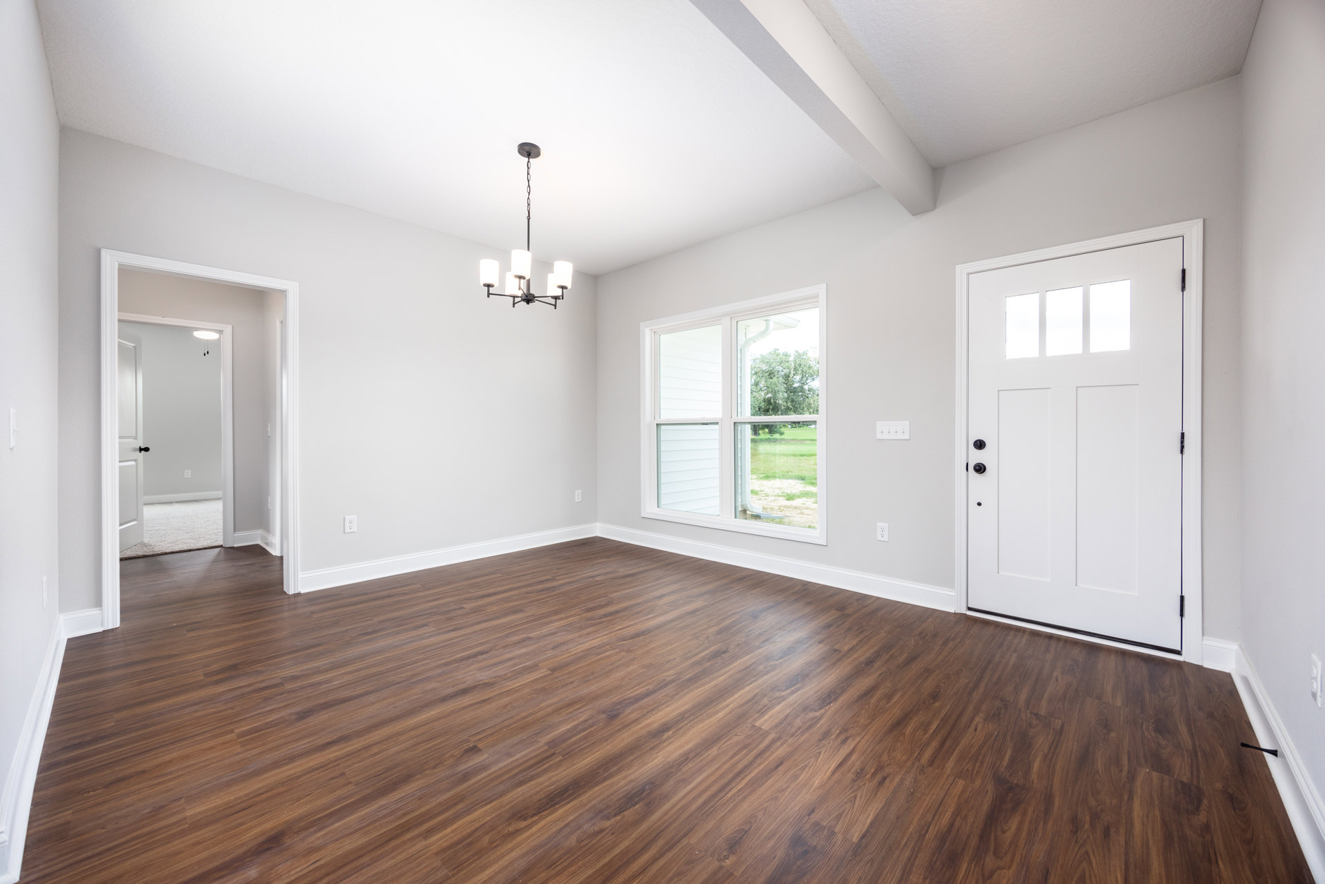 White walls and hardwood floor in a bright room, featuring a white door with black knobs, a modern chandelier, and a window overlooking grass and a gutter.