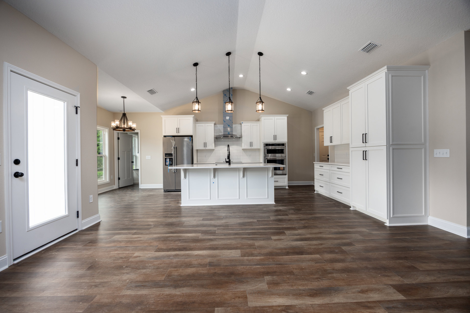 Spacious kitchen featuring white shaker cabinets, wood plank flooring, white island with quartz countertop, stainless steel refrigerator, ceiling vent, and a white door illuminated