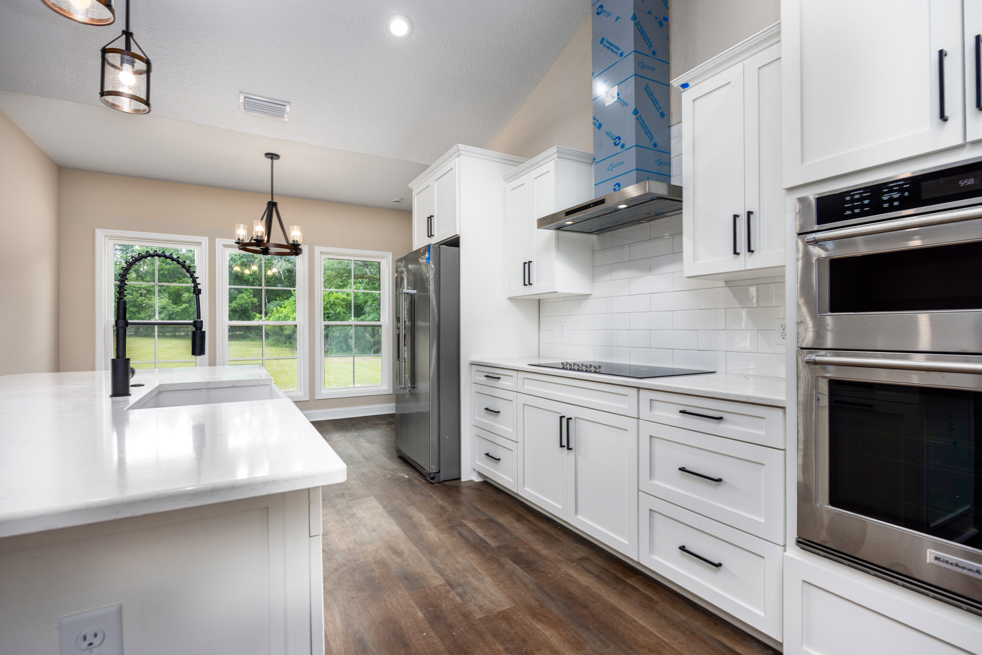 White kitchen with shaker cabinets, stainless steel refrigerator and oven, white countertop, glass pendant light, window with black handle