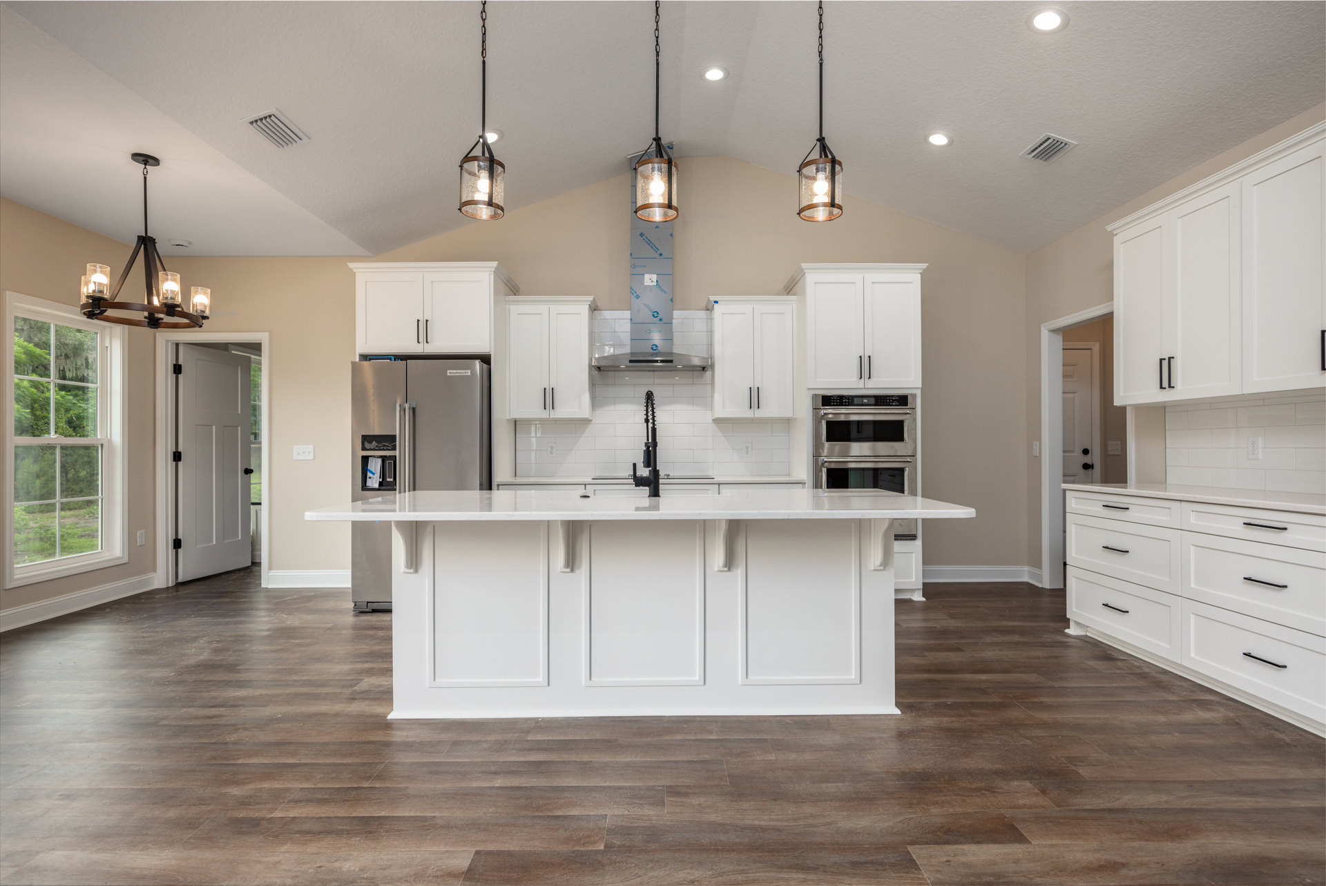 White kitchen featuring a spacious island with light wood base, black faucet at the sink, glass and wood framed pendant lights, large window overlooking trees, and stainless steel