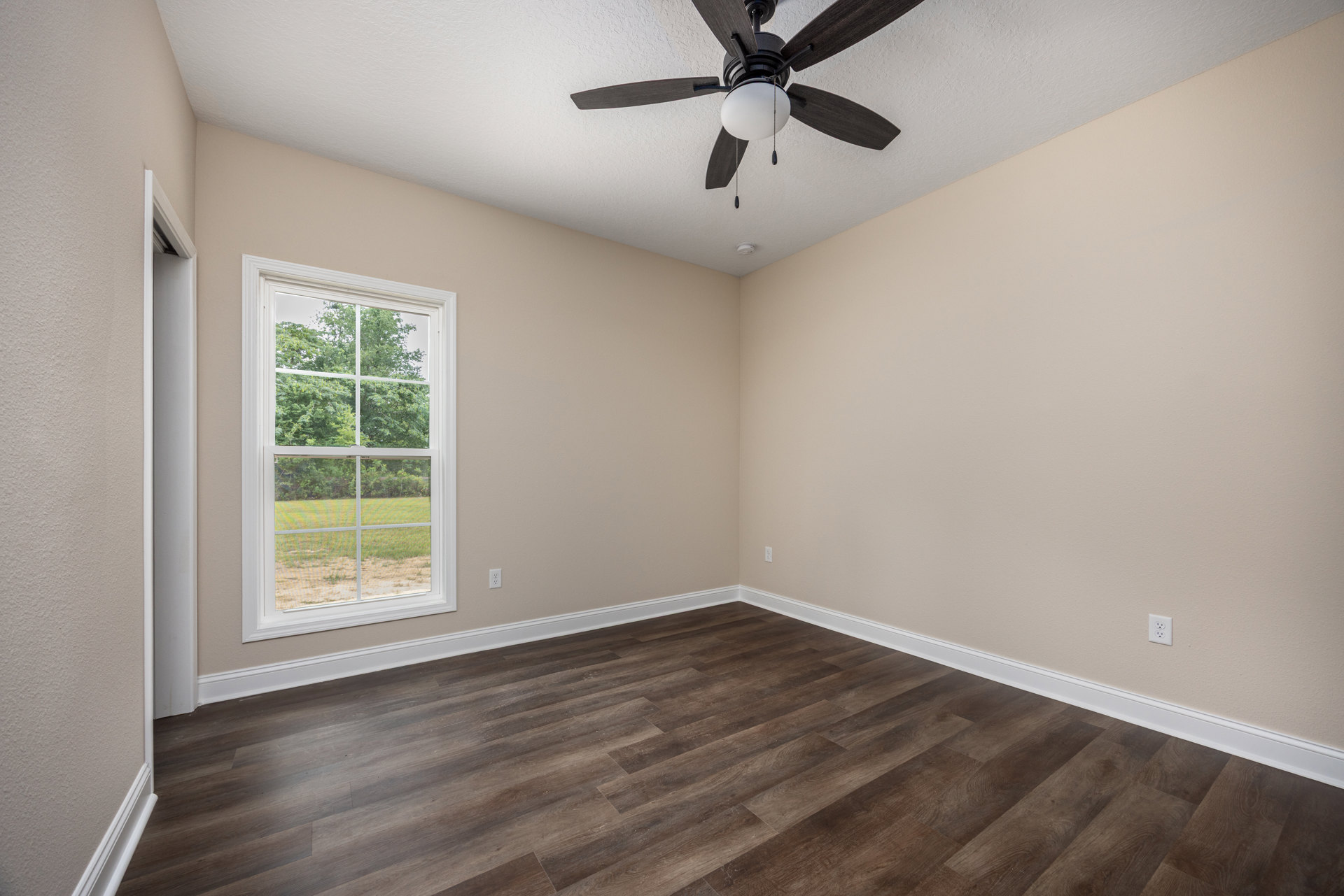 Wood flooring with white baseboards, ceiling fan with light fixture, large window showing trees outside, smooth plaster walls