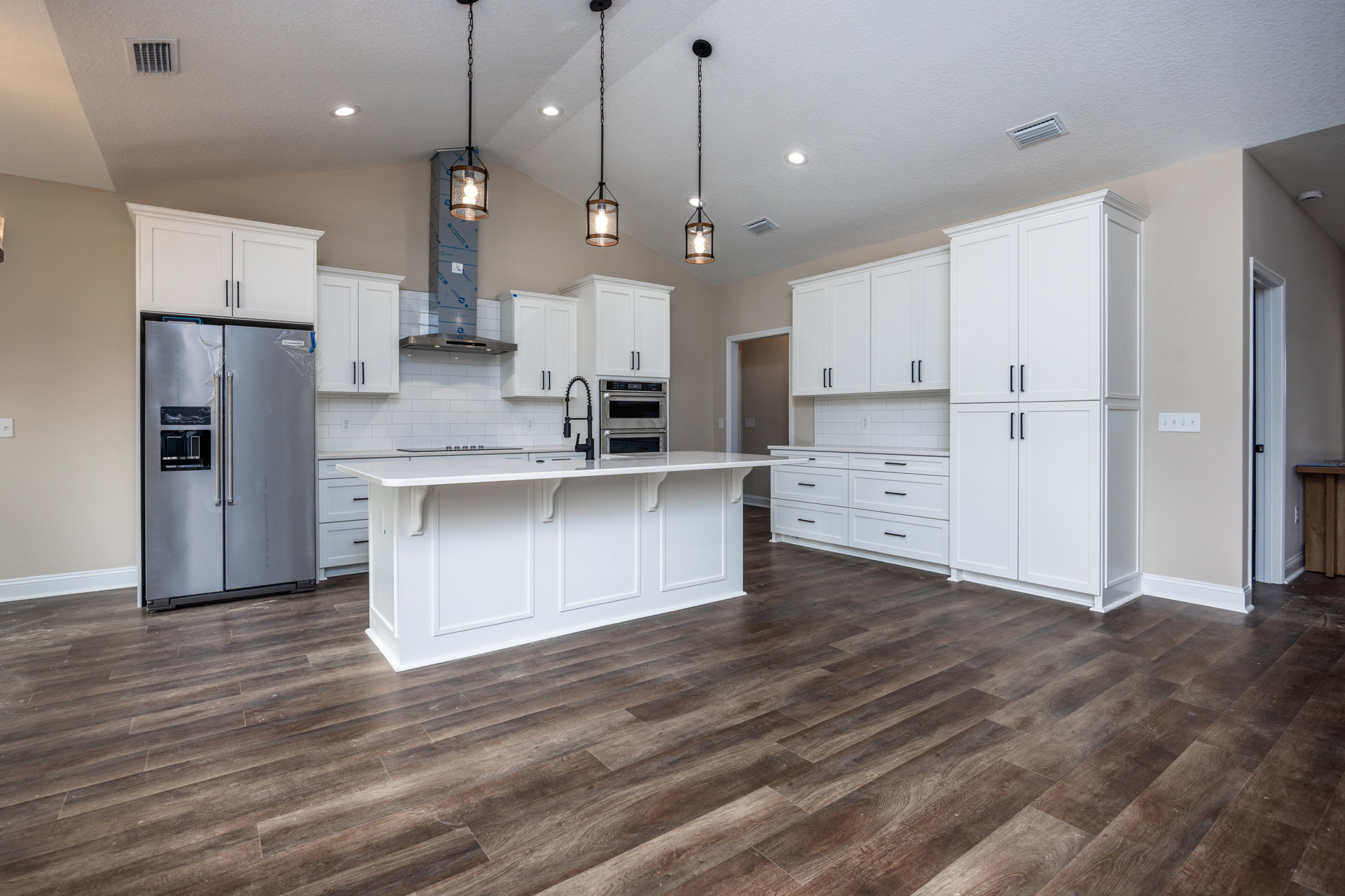 White kitchen with shaker cabinets, wood flooring, stainless steel refrigerator partially wrapped in plastic, built-in oven, black faucet, white trim, and ceiling vent.