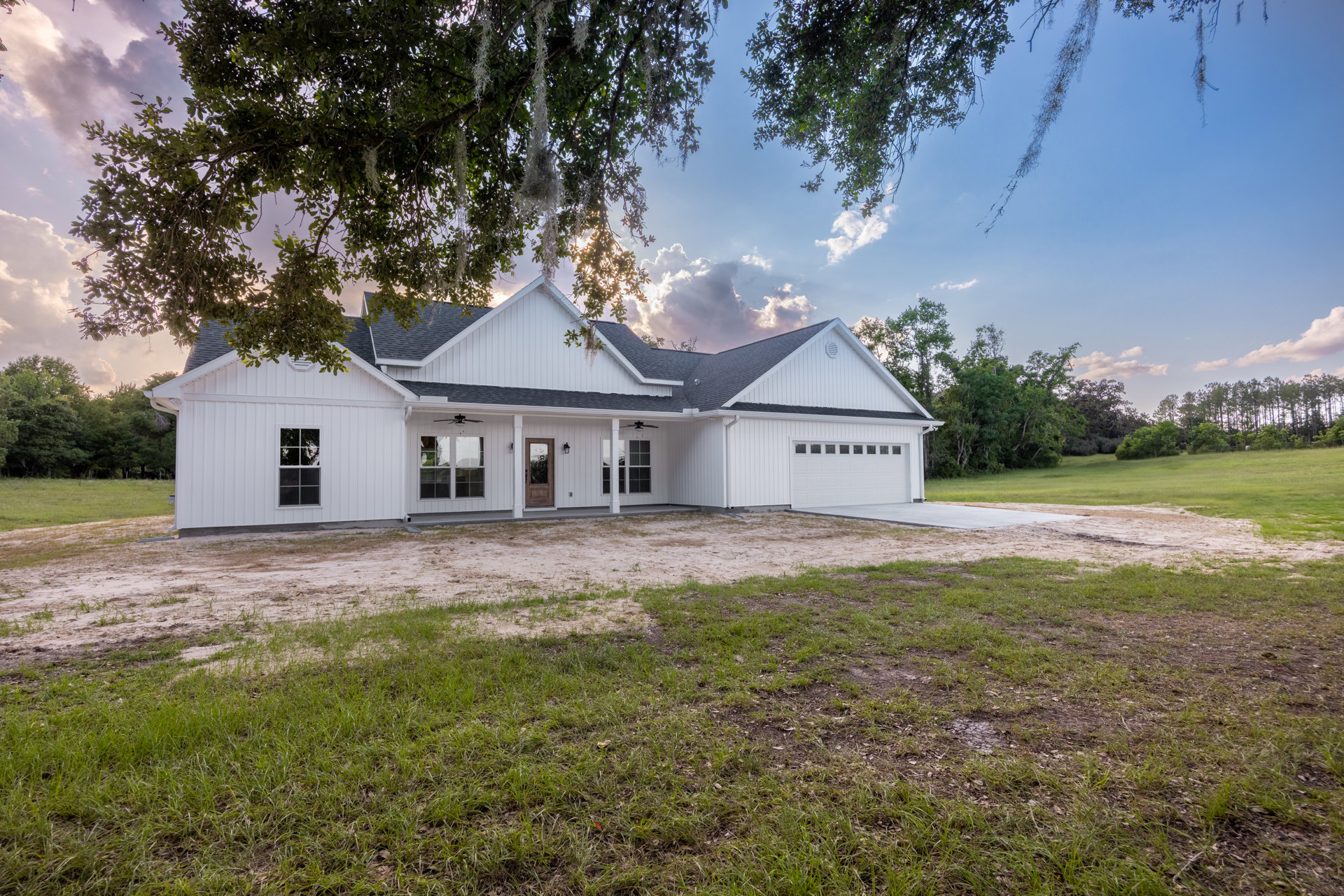 White siding house with brown front door, large paved driveway, green lawn, mature trees, blue sky, multiple windows with white frames