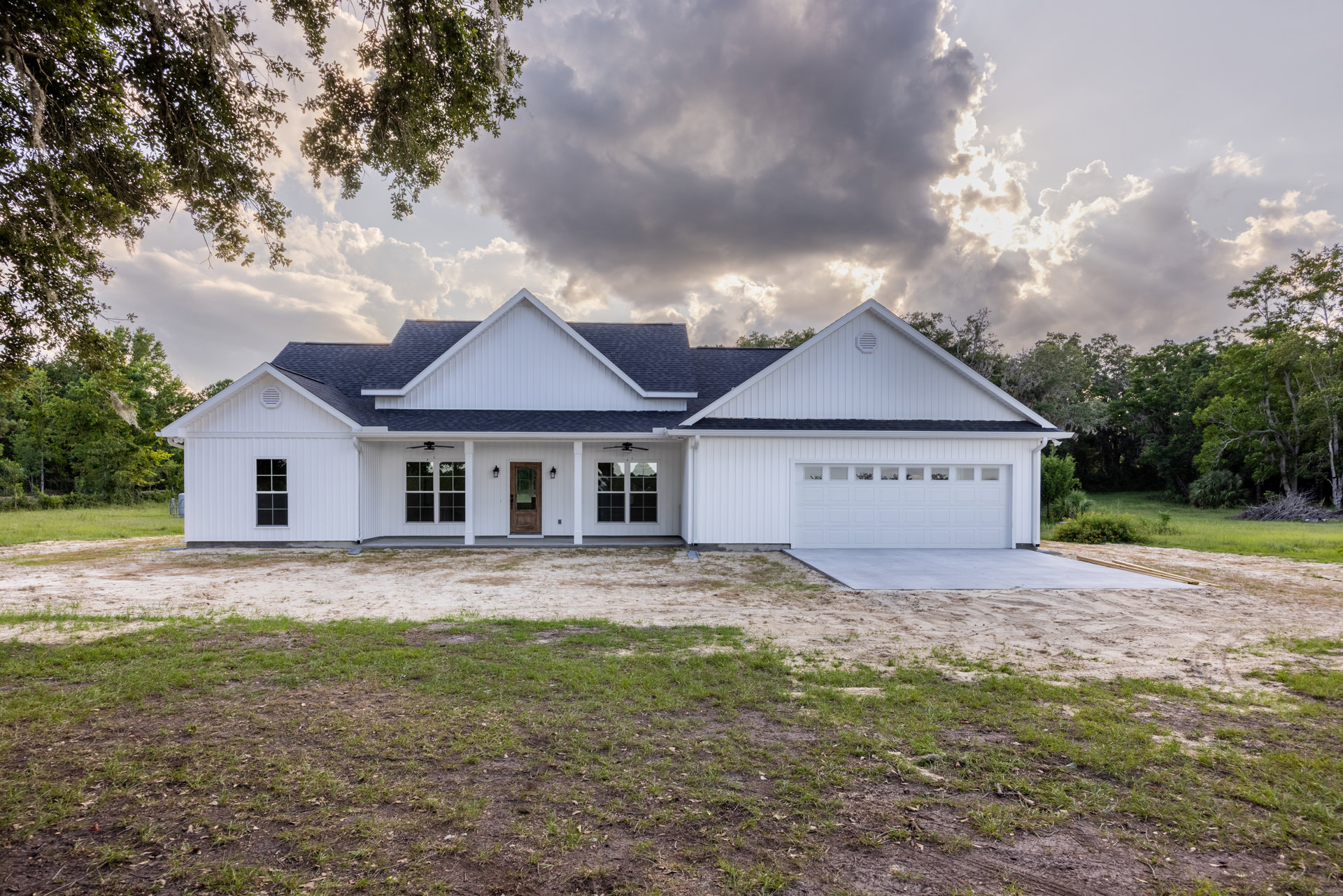 White siding house with black roof, attached garage, concrete driveway, green lawn, mature trees in background, cloudy sky