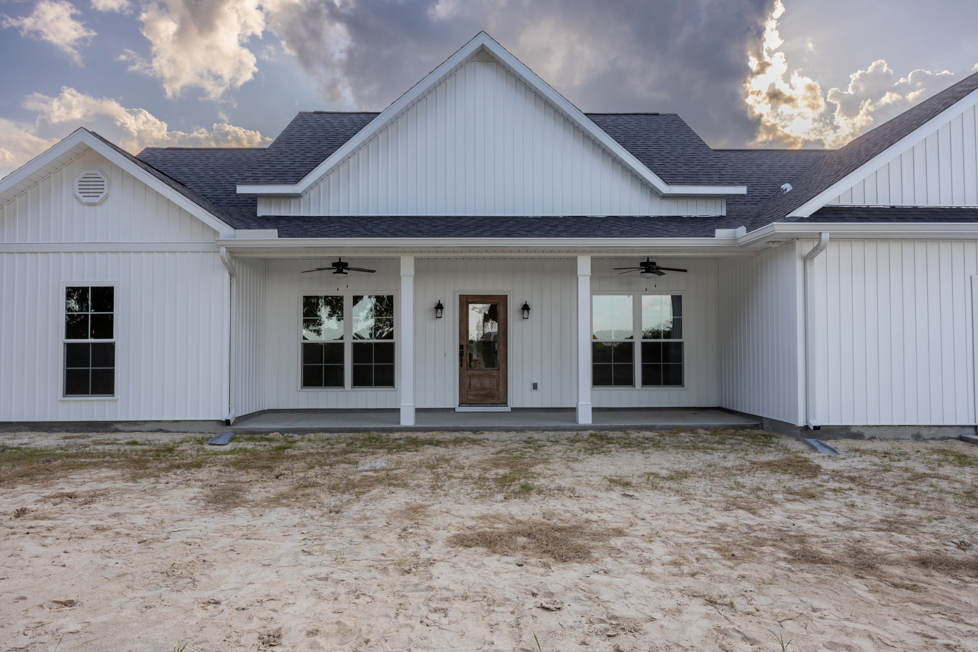 White house with triangular roof, white framed windows, and glass door reflecting a person; dirt and sandy yard with sparse grass.