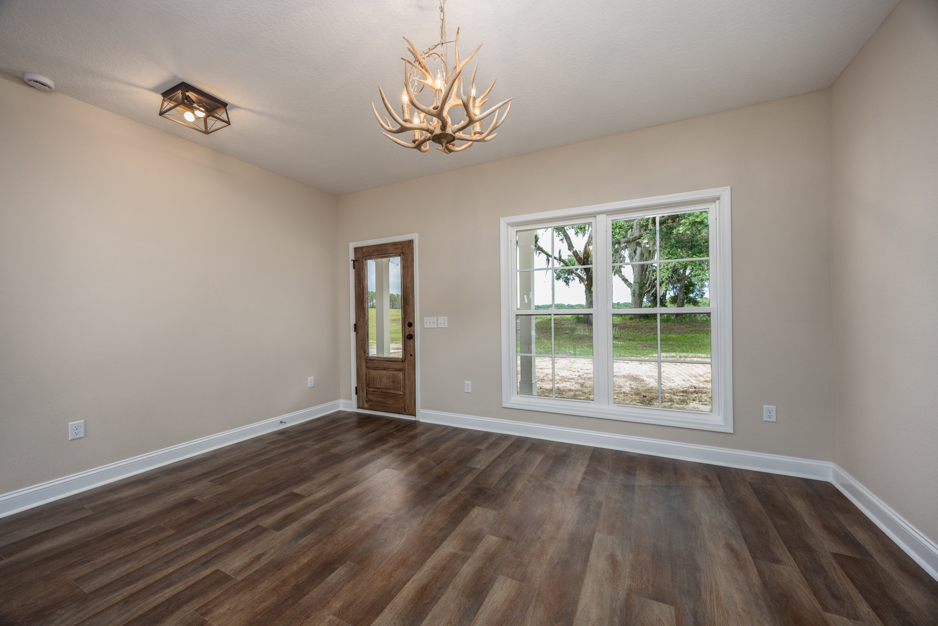 Chandelier with antler design hanging from plaster ceiling, hardwood floor, large window and door with glass panes showing trees and open field outside, white walls with crown