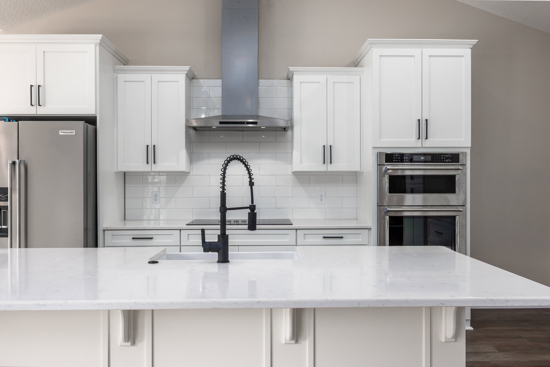 White kitchen with black faucet, white cabinets with black handles, stainless steel oven with glass doors, close-up of refrigerator, white countertop, wooden frame on white wall.
