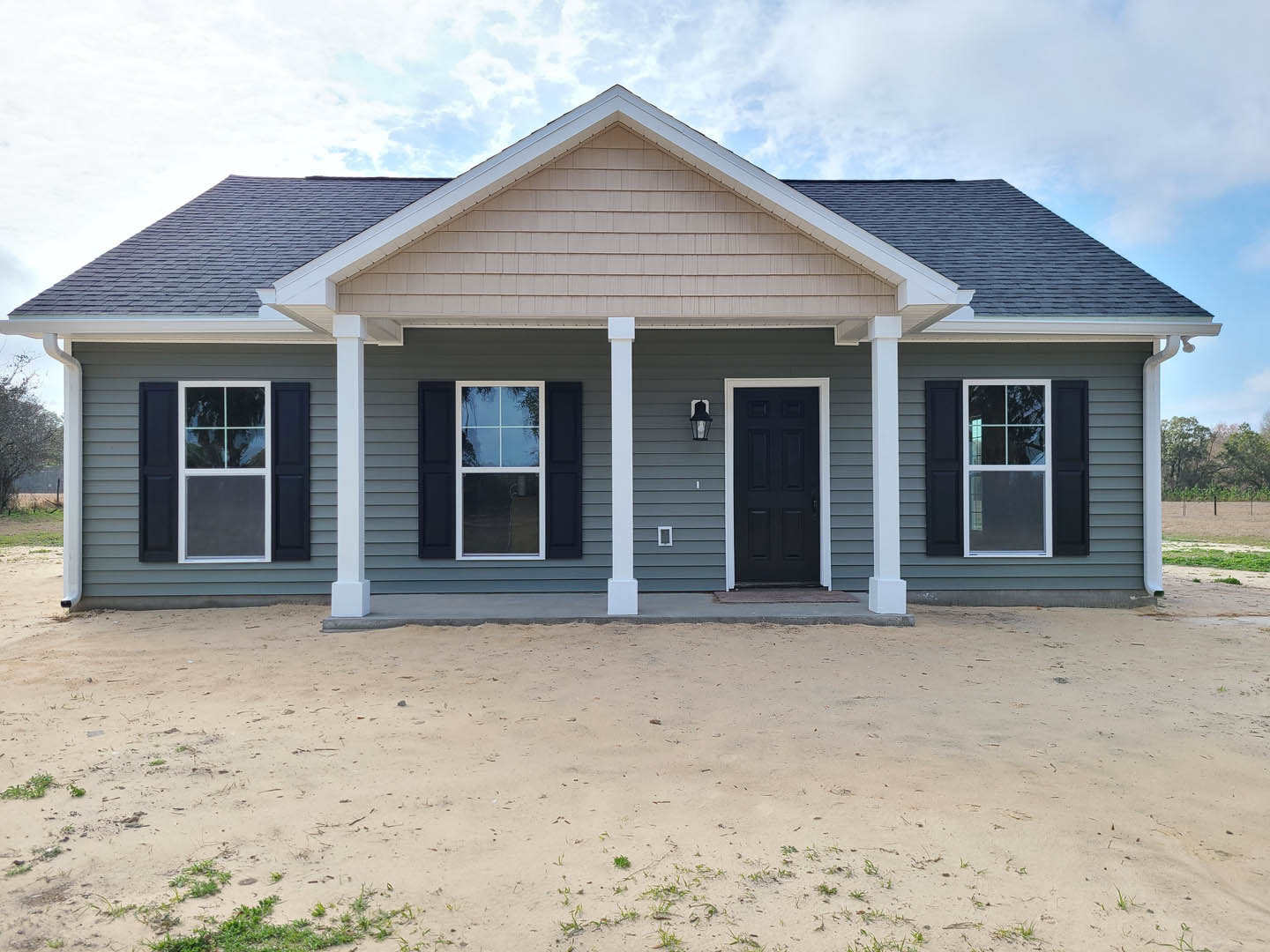 Two-story house with black front door and white trim, grid-style window with white frame, sandy dirt road leading to entrance, patches of grass on ground, cloudy sky overhead