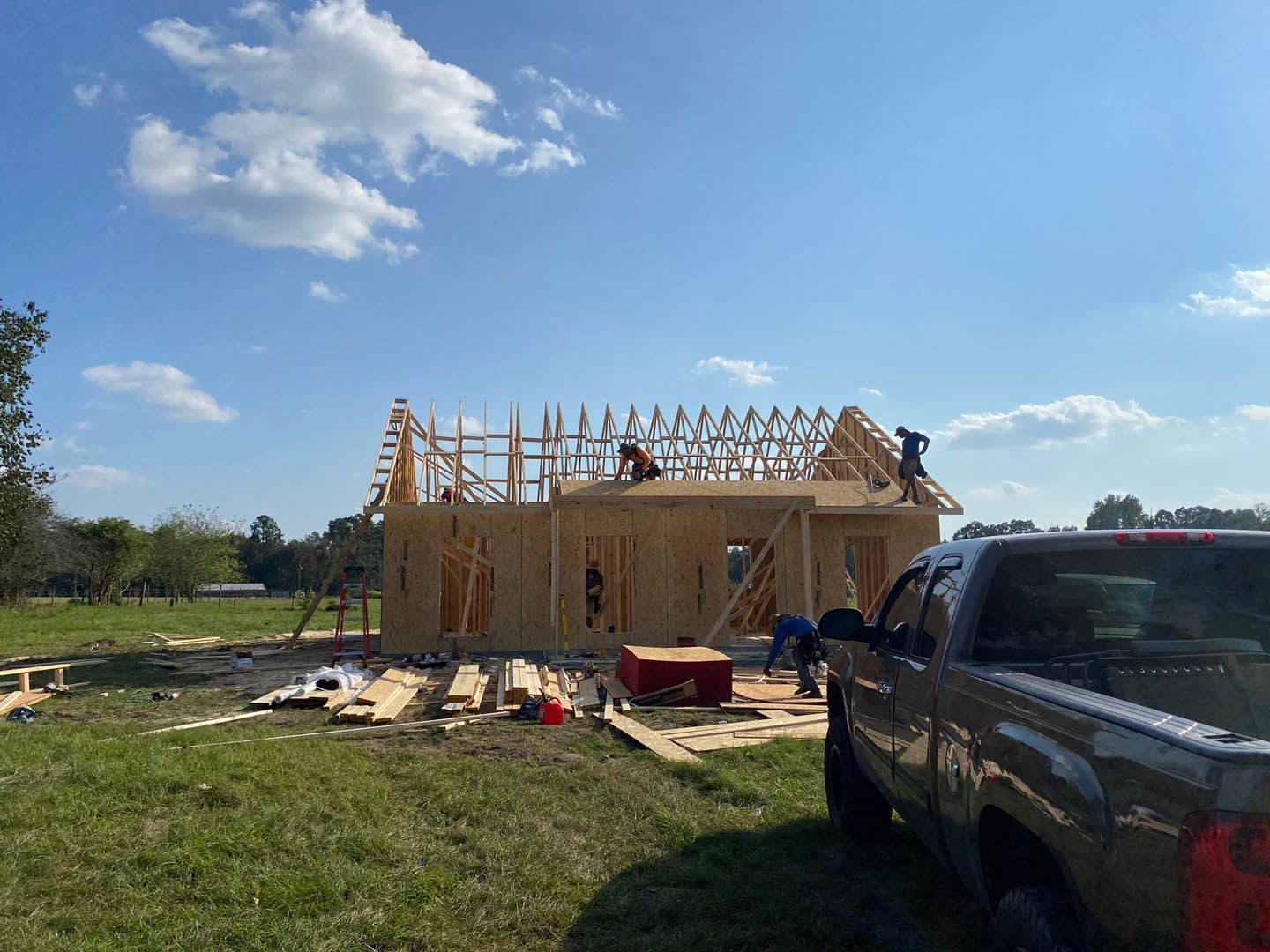 Partially constructed house with exposed wooden framing, worker on roof, truck parked on grassy lawn, car nearby, blue sky with scattered clouds