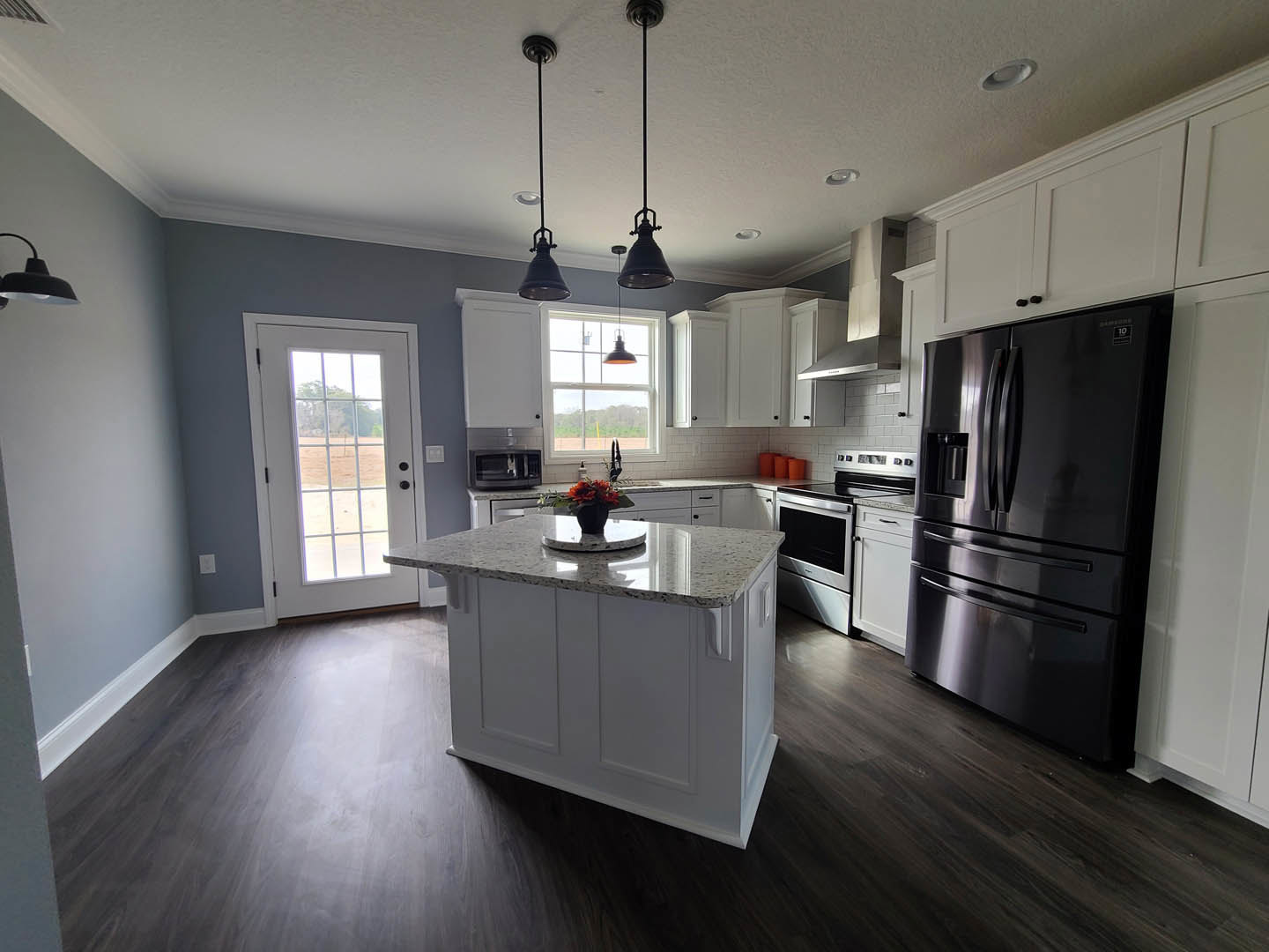 Modern kitchen featuring a two-door black refrigerator, white cabinetry, central island with a flower arrangement, wood flooring, recessed ceiling lights, and a door opening to a