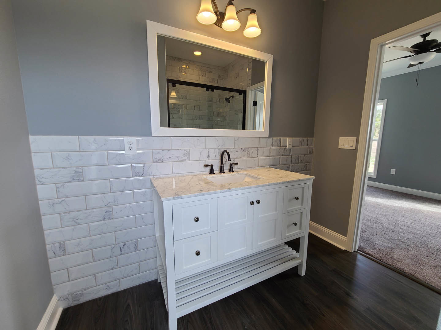 White bathroom vanity with marble countertop, rectangular mirror framed in white, three-light fixture above, chrome faucet, and triple white light switch on pale wall.
